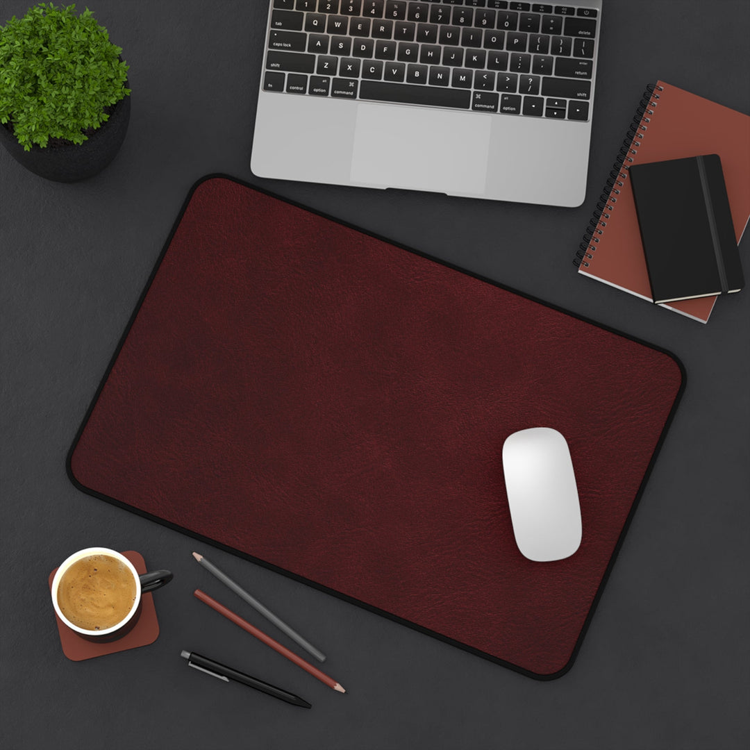 Sunday Theory burgundy leather-look neoprene desk mat with hemmed edges and anti-slip backing on a dark office desk.