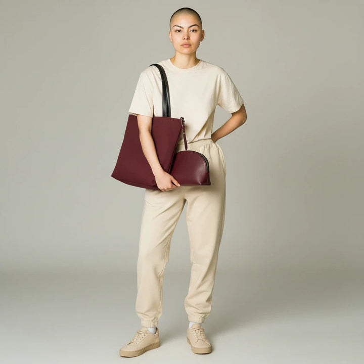 Model holding a Sunday Theory burgundy canvas tote and matching leatherette cosmetic pouch against a neutral backdrop.