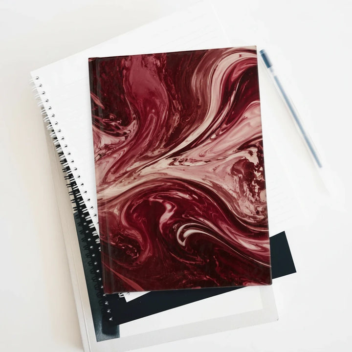 Sunday Theory maroon marble ruled journal with a matte finish, resting on top of spiral notebooks with a pen nearby.