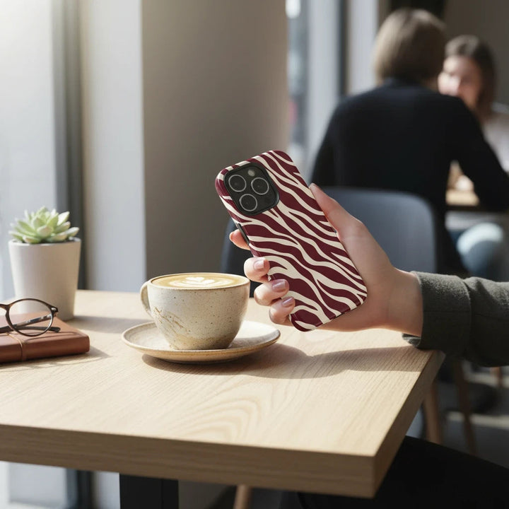 Sunday Theory maroon and cream zebra print protective phone case held in a hand at a cafe table.